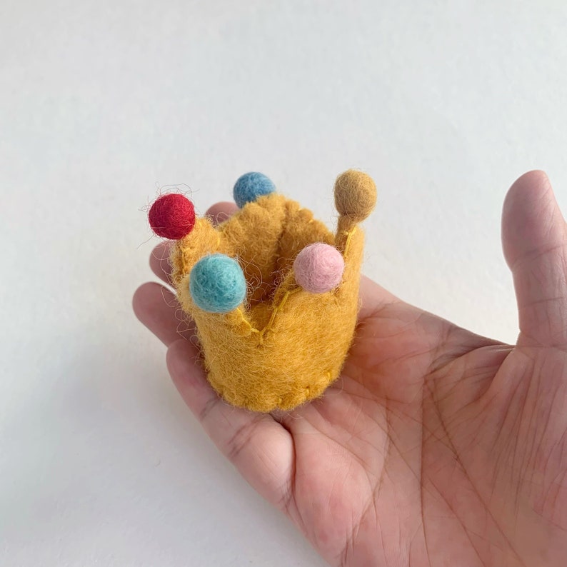 May include: A small, mustard yellow felt crown featuring five points, each topped with a tiny, round, felted ball in red, light blue, dark blue, pink, and tan. The crown is constructed with visible stitching along the edges and base.