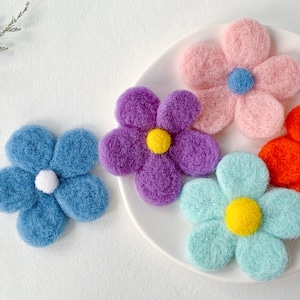 Könnte beinhalten: Fünf Filzblumen-Haarklammern in verschiedenen Farben, darunter Lila, Rosa, Blau und Orange. Jede Blume hat eine weiße Mitte mit einem kleinen Knopf.