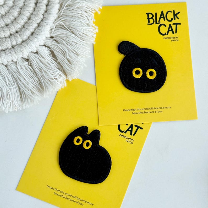 May include: Two black cat embroidery patches with yellow eyes on yellow cards. The cards have the text "BLACK CAT EMBROIDERY PATCH" and "CAT EMBROIDERY PATCH". The cards also have the text "I hope that the world will become more beautiful because of you."
