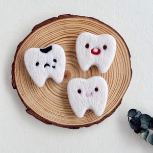 以下が含まれることがあります： 白いフェルト製の歯の形をしたオーナメント3つ。刺繍された顔があり、1つは虫歯があります。丸い木の輪切りに配置されています。歯は異なる表情をしています：悲しい、幸せ、そして驚いた顔。