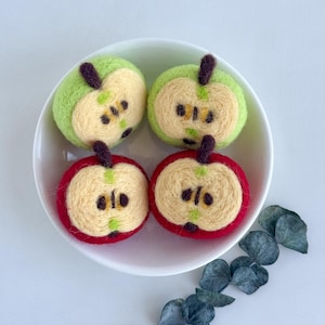 May include: A white ceramic bowl holds four felt apple ornaments. Two are green, and two are red, each cut in half to reveal a beige interior with dark brown seeds. The apples have brown stems and green accents.
