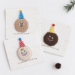 May include: Three embroidered cookie patches on individual white cards labelled cookies Family. One tan cookie with sprinkles and a blue party hat, one dark brown cookie with a yellow party hat, and one white cookie with a red striped party hat.