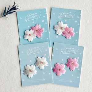 Könnte beinhalten: Vier hellblaue Karten mit gestickten Kirschblüten-Aufnähern. Jede Karte zeigt ein Paar Blüten in Weiß und Rosa oder zwei weiße oder zwei rosa Blüten. Die Karten tragen den Text "Cherry Blossom".