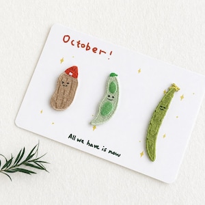 May include: Three embroidered patches featuring a tan peanut wearing a red hat, a light green pea pod, and a long dark green bean, all with smiling faces. The white card displays the text October! and All we have is now with yellow stars.