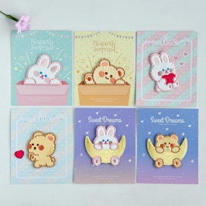 Pode incluir: Seis cartões coloridos com patches bordados que apresentam designs fofos de animais. Os patches incluem um coelho em uma caixa, um urso em uma caixa, um coelho segurando um coração, um urso com um coração, um coelho em uma lua crescente e um urso em uma lua crescente. Os cartões têm texto que diz "Pleasantly Surprised", "Love" e "Sweet Dreams".