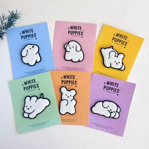 May include: A collection of embroidered puppy patches on pastel-coloured cards. Each patch depicts a white puppy with a black outline. The cards display the text "WHITE PUPPIES" and a short message.