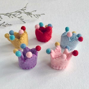 May include: Five small, felted crowns in yellow, red, light blue, purple, and pink. Each crown features a textured, fuzzy surface and is decorated with several tiny, colorful felt balls attached to the points of the crown structure.