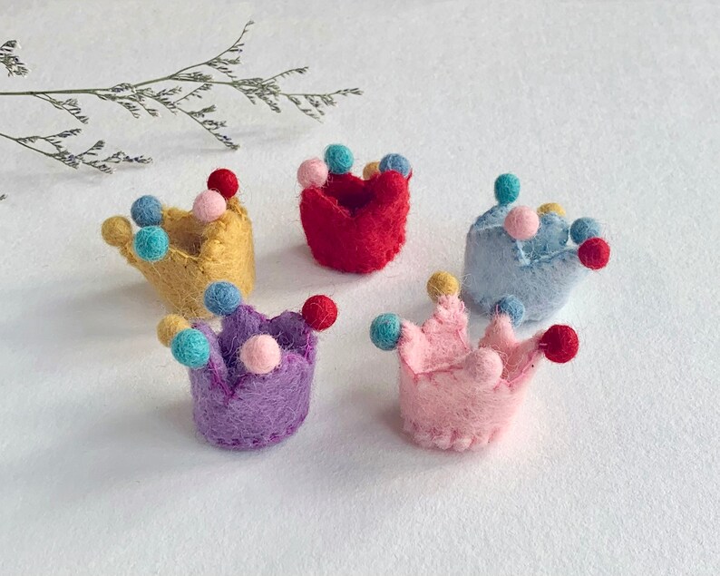 May include: Five small, felted crowns in yellow, red, light blue, purple, and pink. Each crown features a textured, fuzzy surface and is decorated with several tiny, colorful felt balls attached to the points of the crown structure.
