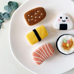 Könnte beinhalten: Ein Set Filz-Essensnadeln mit einem braunen Brot, einer gelben Frühlingsrolle, einer rosa Sushirolle, einer braun-weißen Sushirolle und einem weißen Onigiri mit schwarzem Seetang-Wickel und einem lächelnden Gesicht.