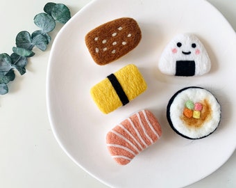 May include: A set of felt food pins featuring a brown bread, a yellow egg roll, a pink sushi roll, a brown and white sushi roll, and a white onigiri with a black seaweed wrap and a smiling face.