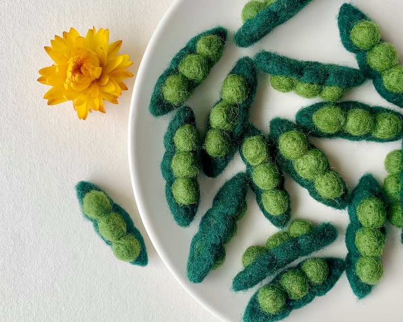 May include: A white plate with green felt pea pods arranged in a decorative pattern. The pea pods are made of felt and have a realistic appearance.