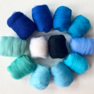 May include: A collection of twelve small, cylindrical bundles of soft, fibrous wool roving arranged in a circular pattern. The colours include various shades of blue, ranging from deep navy and royal blue to light turquoise, sky blue, white, and black.