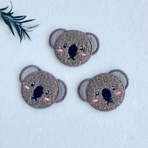 Peut inclure: Trois écussons circulaires en forme de visage de koala avec un fil marron texturé, des nez et des yeux brodés en noir, et de petites joues roses cousues. Chaque écusson possède des oreilles arrondies avec des bordures grises.