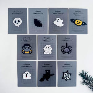 Könnte beinhalten: Eine Sammlung von Halloween-Stickern auf grauen Karten. Die Aufnäher zeigen eine Vielzahl von gruseligen Designs, darunter ein Geist, eine Spinne und ein Kürbis. Jede Karte trägt den weißen Text "Happy Halloween!".