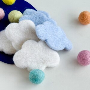 May include: A set of felt cloud shapes in white and light blue, with a variety of felt balls in pink, purple, light green, orange, and teal. The clouds and balls are arranged on a blue background.