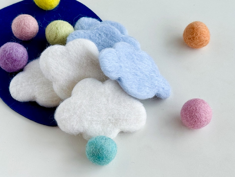 May include: A set of felt cloud shapes in white and light blue, with a variety of felt balls in pink, purple, light green, orange, and teal. The clouds and balls are arranged on a blue background.