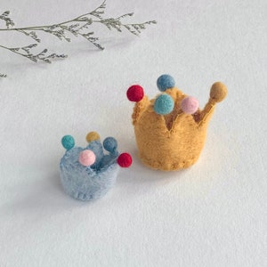 May include: Two small felt crowns, one in light blue and one in mustard yellow, each featuring a series of colorful felt pom-poms in red, light blue, and pink attached to the points. The crowns have a textured, fuzzy appearance and stitched edges.