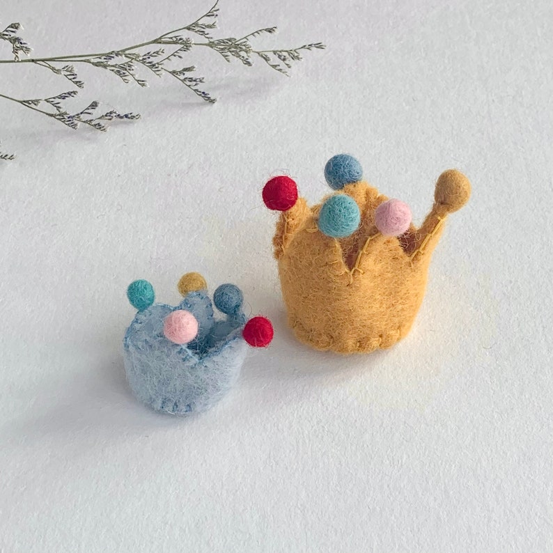 May include: Two small felt crowns, one in light blue and one in mustard yellow, each featuring a series of colorful felt pom-poms in red, light blue, and pink attached to the points. The crowns have a textured, fuzzy appearance and stitched edges.