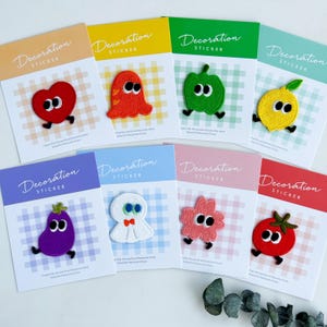 May include: A selection of decorative stickers with embroidered designs: a red heart, orange ghost, green apple, yellow lemon, purple aubergine, white cloud, pink flower, and red tomato. Each sticker is on a card with the text "Decoration STICKER".