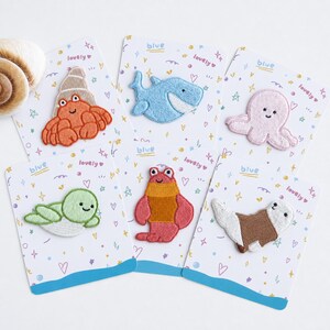 May include: Six embroidered iron-on patches featuring sea creatures on individual white backing cards. The designs include an orange hermit crab, a light blue shark, a pink octopus, a light green sea turtle, a red lobster, and a brown seal.