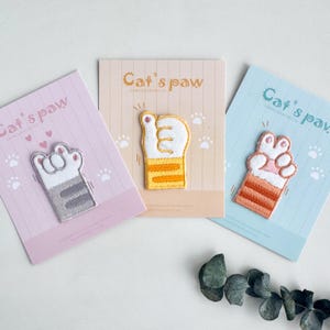 以下が含まれることがあります： カードに刺繍された3つの猫の足跡パッチ。パッチは白、グレー、オレンジ、ピンクです。カードはピンク、ベージュ、青で、「Cat's paw」の文字と足跡が描かれています。