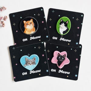 May include: Four embroidered cat patches on black square display cards with star patterns and the text OH Meow. The patches feature an orange cat in a circle, a black and white cat in an oval, a grey cat in a heart, and a black cat in a star shape.