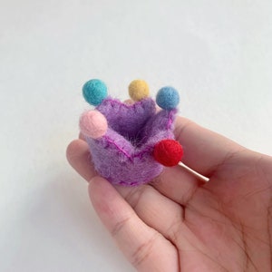 May include: A small, purple felt crown featuring five points, each topped with a tiny, solid-colored felt ball in shades of light blue, yellow, pink, and red. The crown is constructed from fuzzy, textured wool material with visible pink stitching.