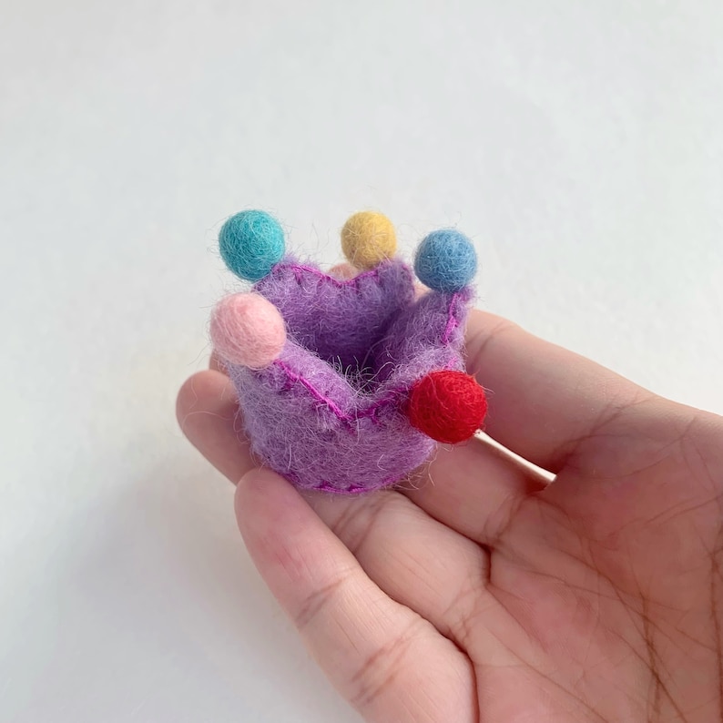 May include: A small, purple felt crown featuring five points, each topped with a tiny, solid-colored felt ball in shades of light blue, yellow, pink, and red. The crown is constructed from fuzzy, textured wool material with visible pink stitching.