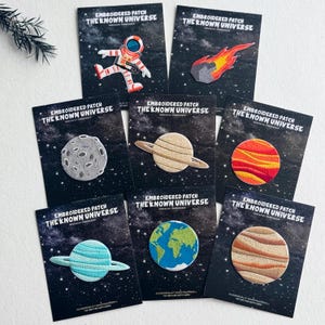 Könnte beinhalten: Acht gestickte Aufnäher mit Weltraummotiven, darunter ein Astronaut, ein Komet und verschiedene Planeten. Jeder Aufnäher ist auf einer schwarzen Karte mit der Aufschrift Embroidered Patch The Known Universe und Sternenhimmel-Design befestigt.