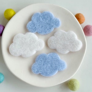 May include: Four felt clouds in different shades of blue and white. The clouds are arranged in a circle on a white plate.