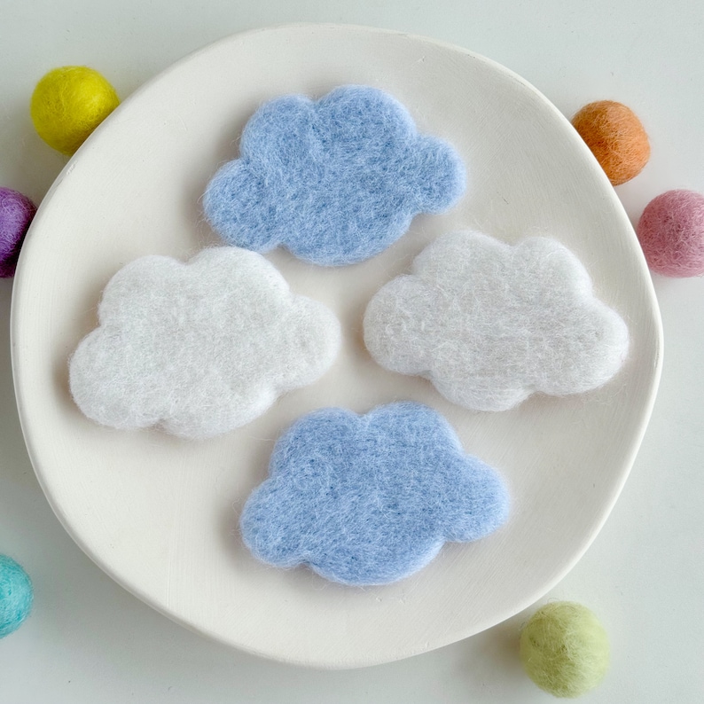 May include: Four felt clouds in different shades of blue and white. The clouds are arranged in a circle on a white plate.