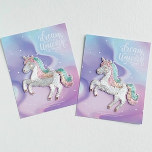 Könnte beinhalten: Zwei weiße Einhorn-Bügeleisen-Patches mit rosa, grünen und goldenen Akzenten auf einem lila und blauen Hintergrund. Die Patches sagen "dream like a unicorn" in weißer Schrift.