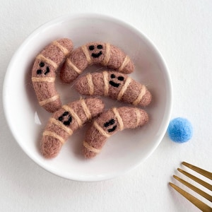 Pode incluir: Uma tigela de cerâmica branca contém cinco enfeites de feltro em forma de croissant sorridentes. Os enfeites são de cor rosa empoeirado com listras bege e rostos bordados em preto. Um pequeno pompom azul e um garfo dourado também estão na imagem.