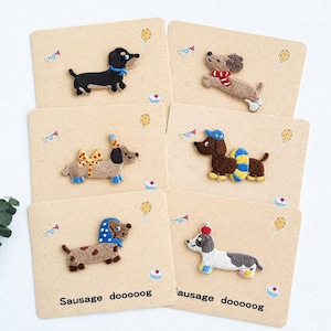 Könnte beinhalten: Sechs bestickte Dackel-Aufnäher auf einzelnen beigen Karten. Jeder Aufnäher zeigt ein anderes Hundedesign mit Accessoires wie Schals, Hüten oder Schwimmringen. Die Karten sind mit dem Text Sausage dooooog, Luftballons und Cupcakes bedruckt.