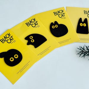 May include: Four yellow cards with black cat patches. Each patch has a different cat shape with yellow embroidered eyes. The cards read "BLACK CAT EMBROIDERY PATCH" in black text. A sprig of greenery is in the background.