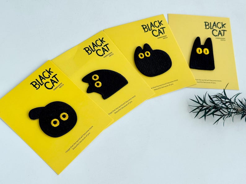May include: Four yellow cards with black cat patches. Each patch has a different cat shape with yellow embroidered eyes. The cards read "BLACK CAT EMBROIDERY PATCH" in black text. A sprig of greenery is in the background.