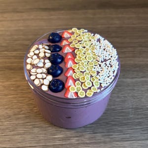 May include: A clear plastic container filled with purple slime topped with decorative clay pieces shaped like blueberries, strawberry slices, kiwi slices, and small white sprinkles arranged in distinct rows across the surface.