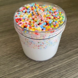 Pode incluir: Um recipiente de plástico transparente cheio de slime branco e contas de espuma coloridas. As contas são de várias cores, incluindo vermelho, amarelo, azul e verde. O recipiente está sobre uma superfície de madeira.