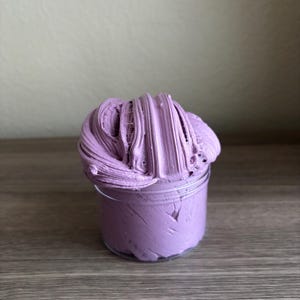 May include: A clear plastic cylindrical container filled with a thick, textured lavender-colored substance. The material has a dense, whipped consistency with visible ridges and folds that extend above the rim of the container in a mound.