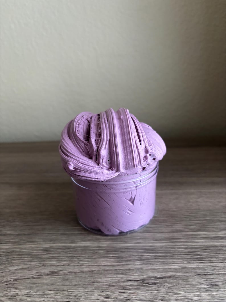 May include: A clear plastic cylindrical container filled with a thick, textured lavender-colored substance. The material has a dense, whipped consistency with visible ridges and folds that extend above the rim of the container in a mound.