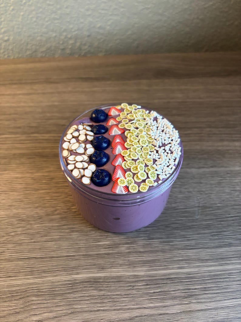 May include: A clear plastic container filled with purple slime topped with decorative clay pieces shaped like blueberries, strawberry slices, kiwi slices, and small white sprinkles arranged in distinct rows across the surface.