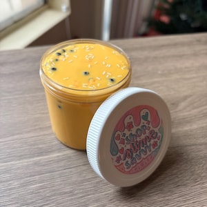 May include: A clear plastic container filled with yellow slime, speckled with small black and white pieces. The lid, off to the side, has a white rim and a pink, blue, and red graphic that reads "Shop Wine Savvy".