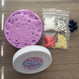 May include: A round container filled with purple slime, a white lid featuring a sticker that reads SHOP SLIME SAVVY, and four small plastic bags containing white sprinkles, brown and white slices, red heart shapes, and yellow fruit slices, plus five dark blue beads.