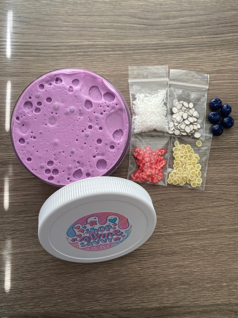 May include: A round container filled with purple slime, a white lid featuring a sticker that reads SHOP SLIME SAVVY, and four small plastic bags containing white sprinkles, brown and white slices, red heart shapes, and yellow fruit slices, plus five dark blue beads.