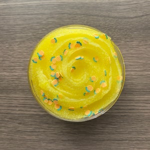 May include: A clear plastic container filled with bright yellow slime. The slime is decorated with small, colorful pineapple-shaped confetti. The background is a wooden surface.