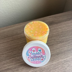 Pode incluir: Um pote de plástico transparente cheio de slime amarelo contendo pequenos pingentes coloridos em forma de fruta. A tampa branca apresenta um adesivo circular com um design de gotejamento pastel e o texto SHOP SLIME SAVVY cercado por ícones.