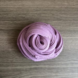 May include: A mound of lavender-colored slime with a swirled, textured surface. The substance is shaped into a dense, coiled mass, displaying a consistent light purple hue throughout its pliable, folded layers and smooth, rounded ridges.