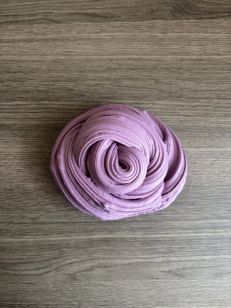 May include: A mound of lavender-colored slime with a swirled, textured surface. The substance is shaped into a dense, coiled mass, displaying a consistent light purple hue throughout its pliable, folded layers and smooth, rounded ridges.