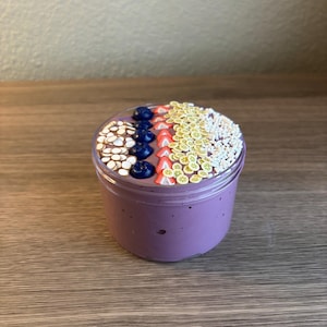 May include: A clear plastic jar filled with purple slime topped with decorative polymer clay slices. The toppings include rows of white almond shapes, dark blue blueberries, red strawberry slices, yellow banana coins, and small white sprinkles.
