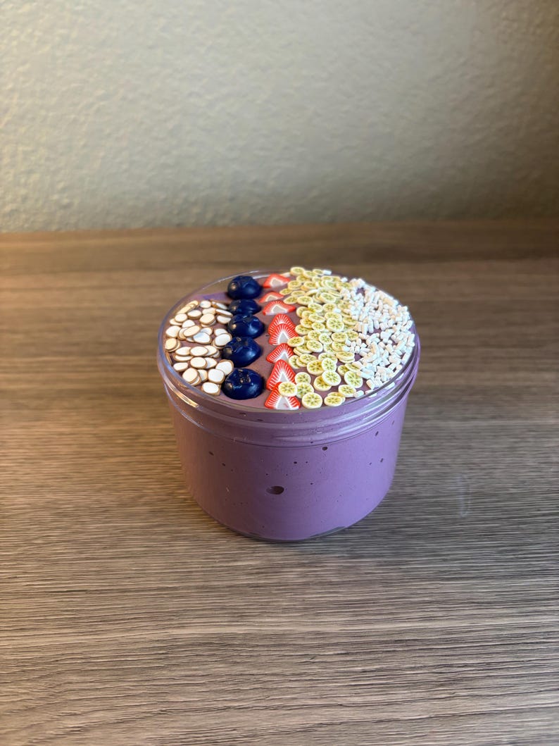 May include: A clear plastic jar filled with purple slime topped with decorative polymer clay slices. The toppings include rows of white almond shapes, dark blue blueberries, red strawberry slices, yellow banana coins, and small white sprinkles.
